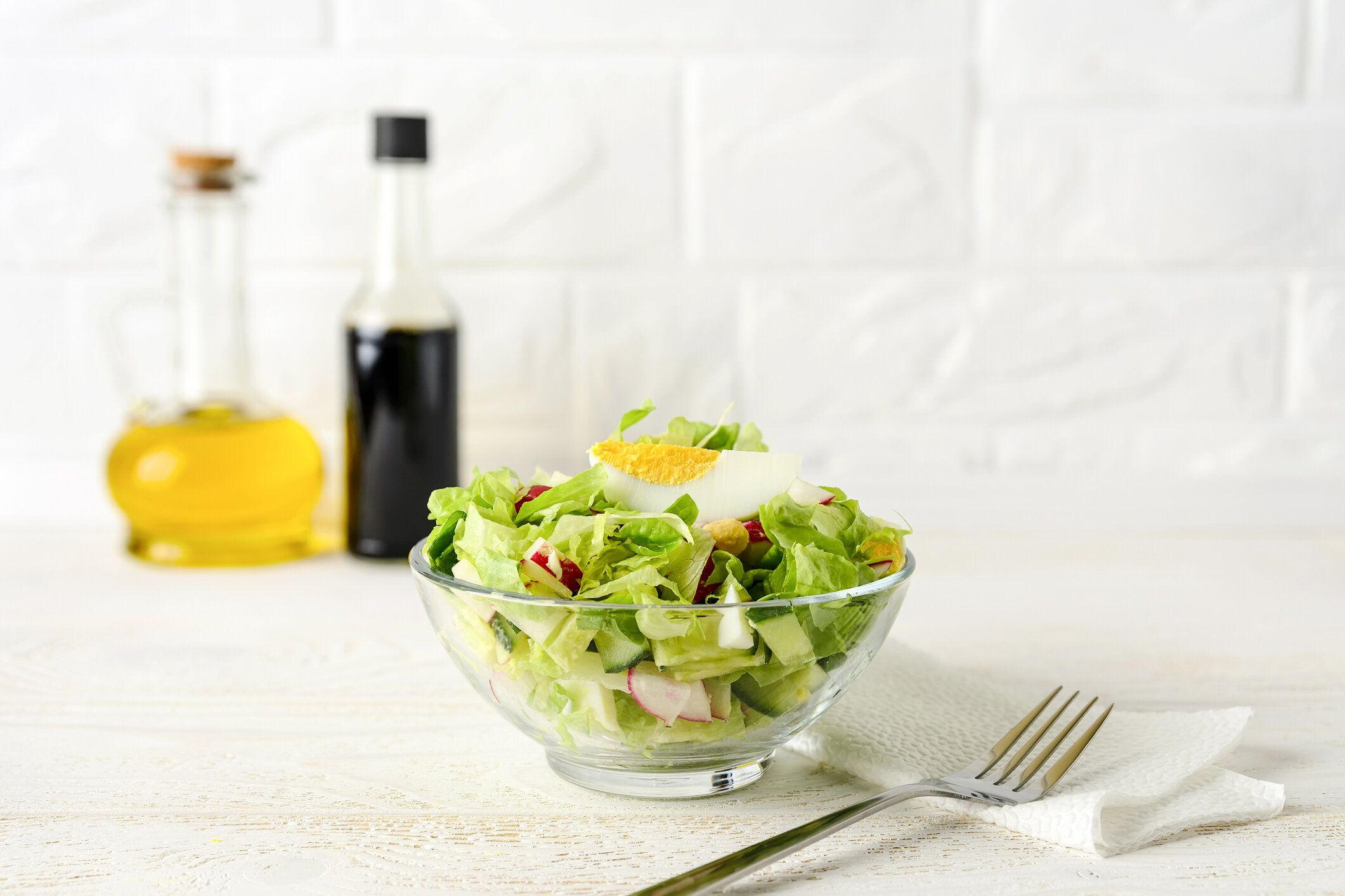 白い木のテーブルの上にガラスボウルにレタス、キュウリ、大根、ゆで卵の新鮮な自家製サラダ。背景にドレッシングのためのオリーブオイルと醤油。健康的な食事。ハイキーショット。