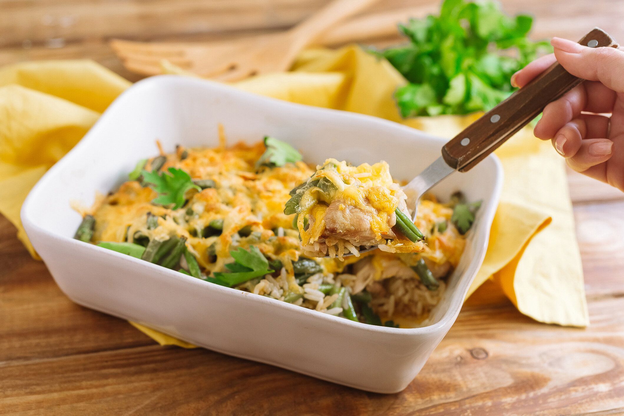 チーズクラストと醤油と米と焼き鶏もも肉のフィレ。長粒米とグリーンビーンズ。木製のテーブルの上に白いベーキングディッシュキャセロール、黄色の生地ナプキン、パセリの葉。