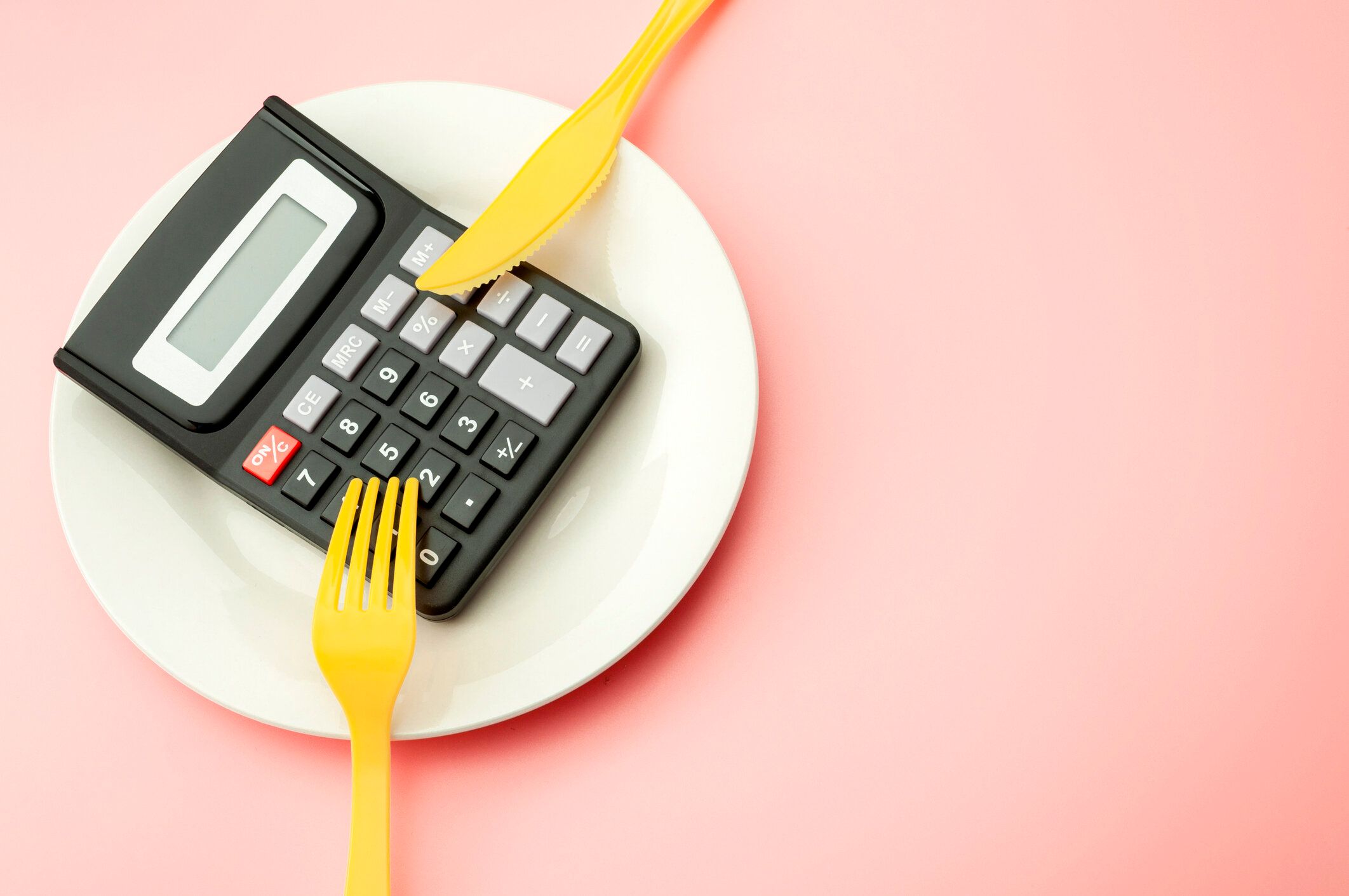 高価な食品支出コストを計算し、空のプレート上の電卓、黄色のフォーク、ナイフをコピースペースでピンクの背景に隔離してカロリーと減量プログラムの概念を数えます