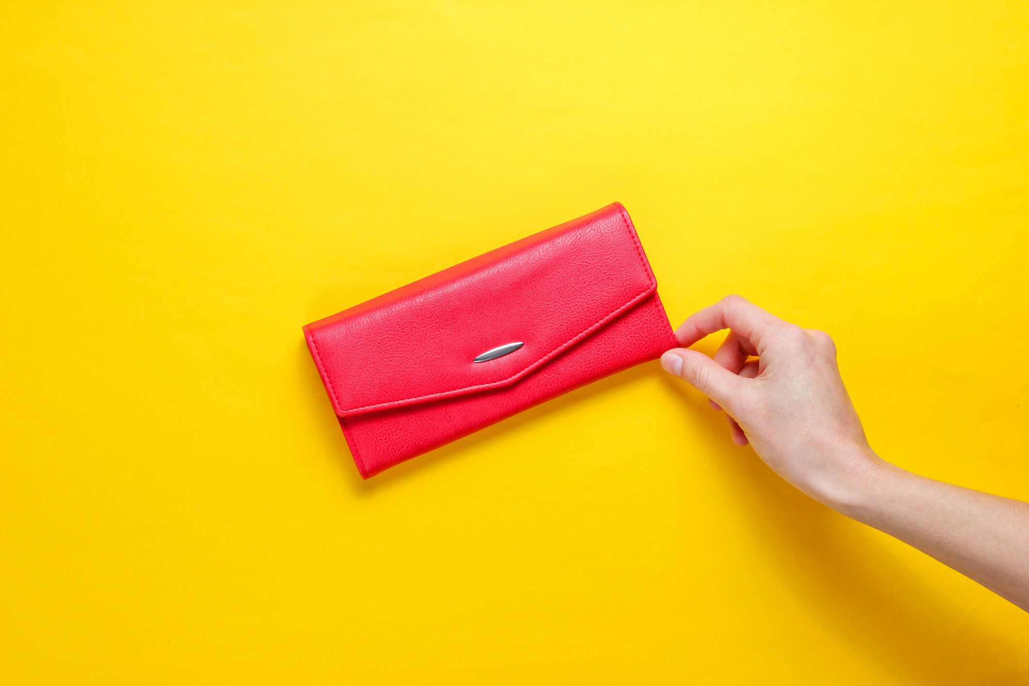 ミニマリズムトレンド。女性の手は、黄色の背景に赤い革の財布を取ります。トップビュー