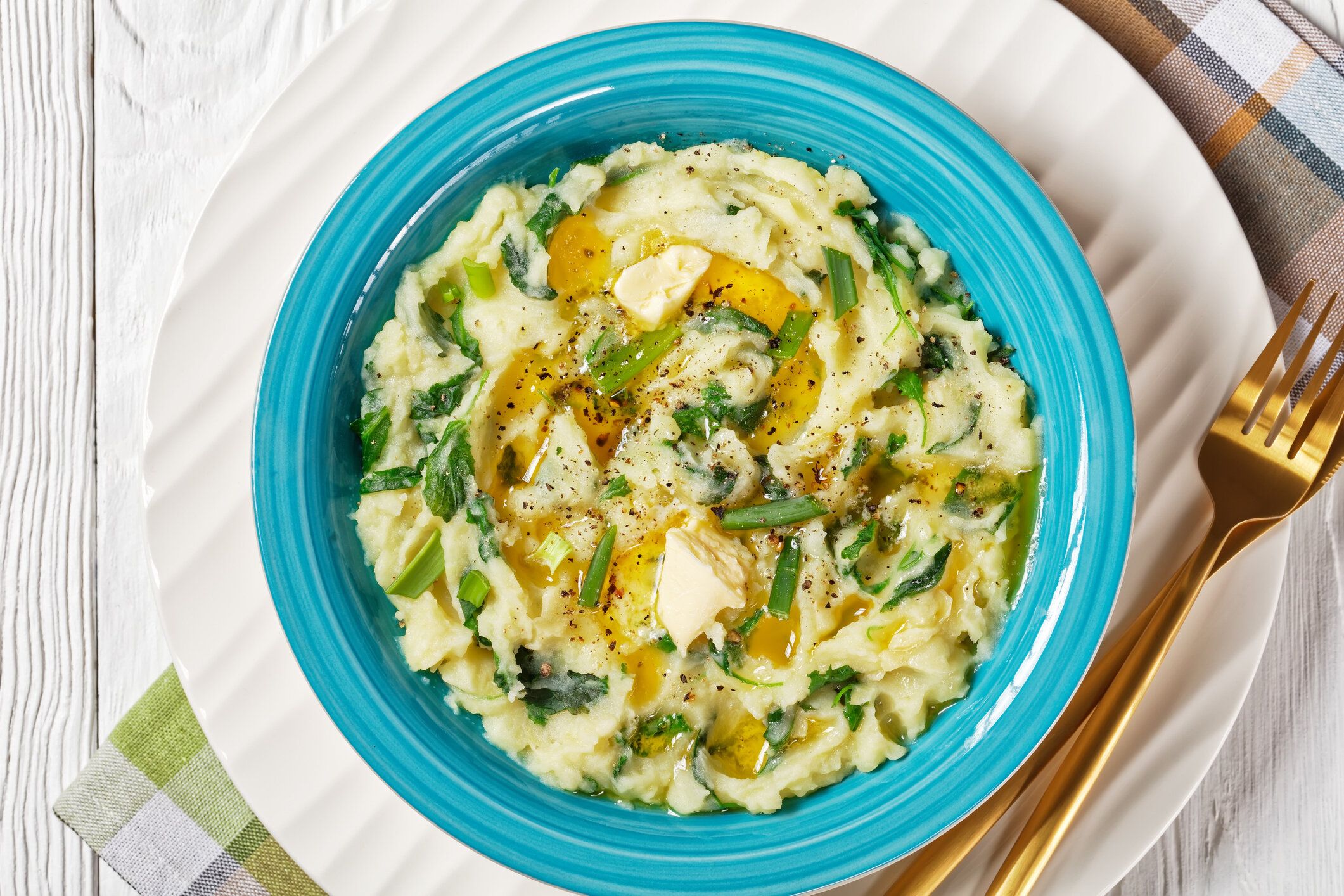 アイルランドのコルキャノン伝統的なポテトマッシュとケール、ニンニク、溶かしたバター、ネギは、白い木製の背景にカトラリー、トップビュー、フラットレイ、クローズアップで青いボ�