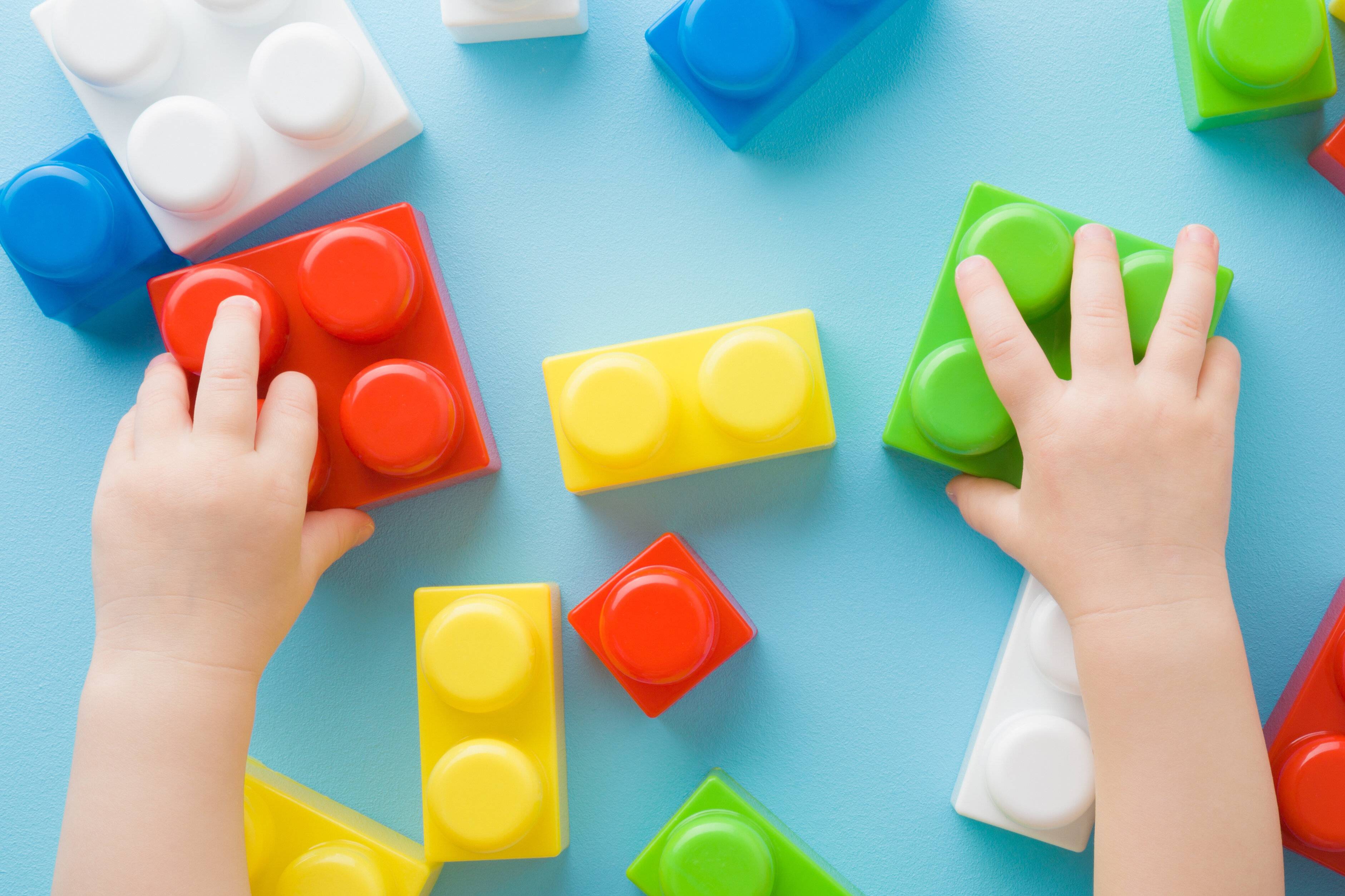 赤ちゃんの女の子の手は、水色のテーブルの背景にカラフルなプラスチックブロックで遊んでいます。クローズ アップ。小さな子供のための開発のおもちゃ。視点ショット。トップダウンビ