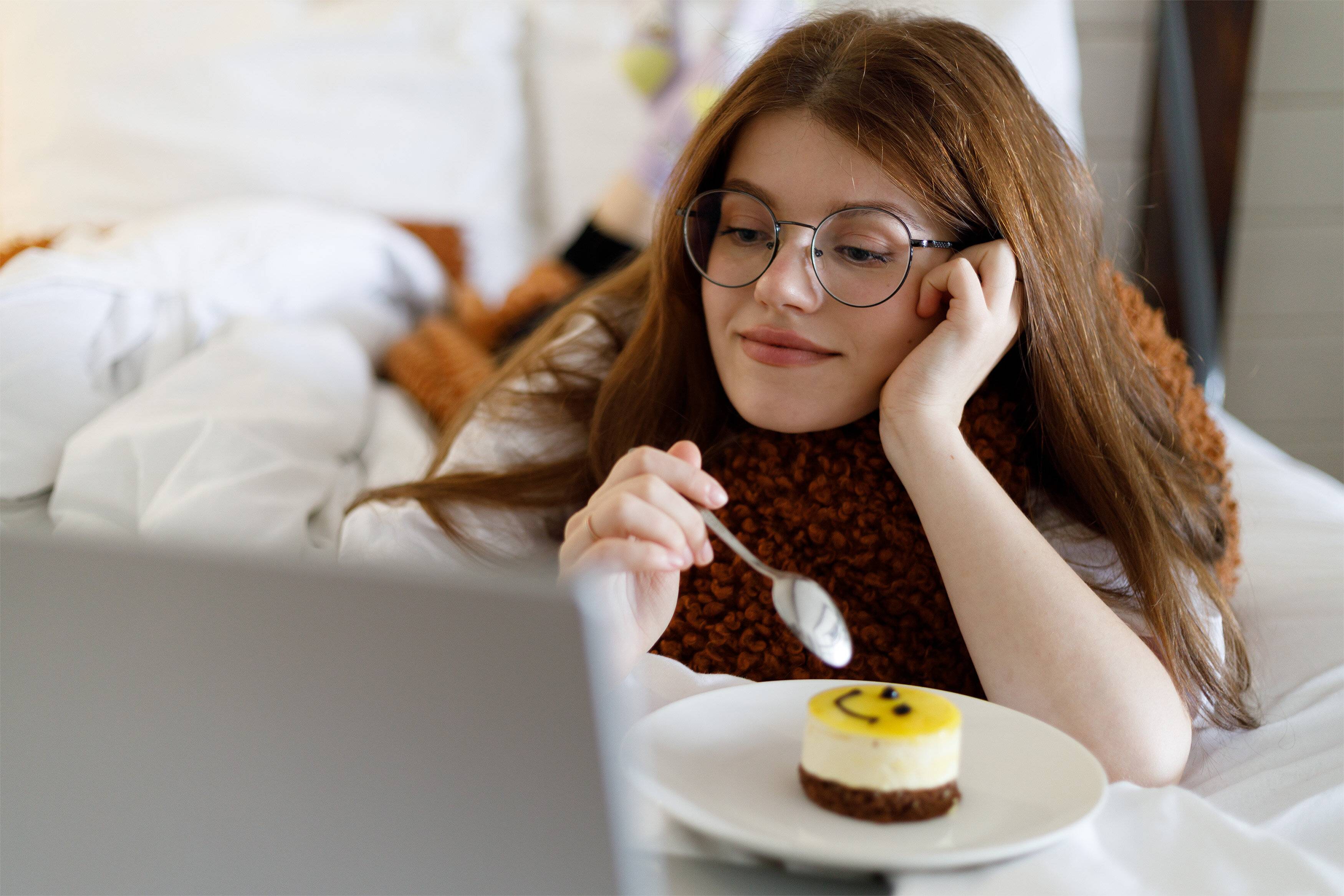 美しい若い女性がラップトップを持ってベッドに横たわり、映画を見たり、お菓子を食べたりしています。