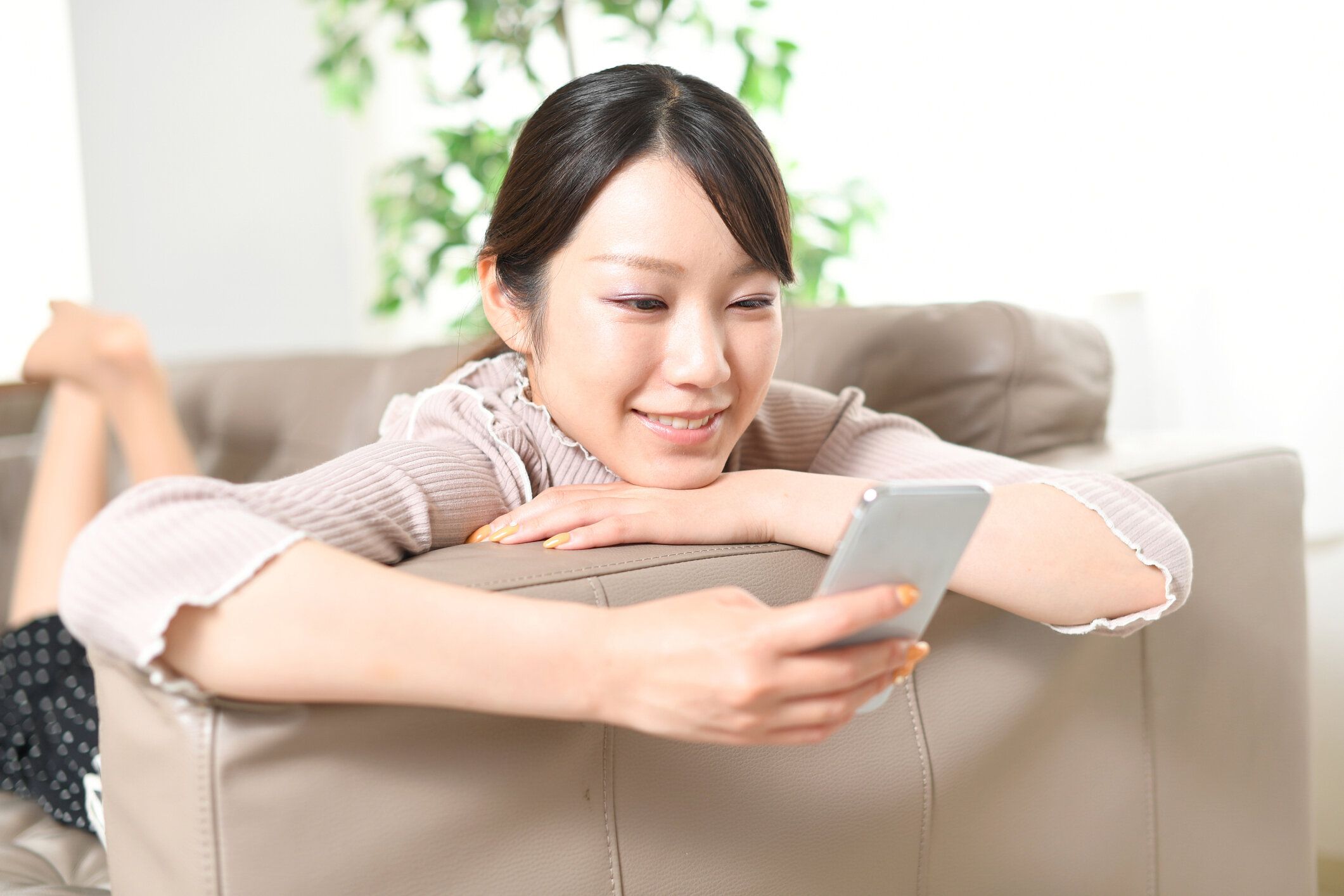 カジュアルな服を着たアジアの若い女性が、ソファに横たわりながら笑顔でスマートフォン(携帯電話)を見ている