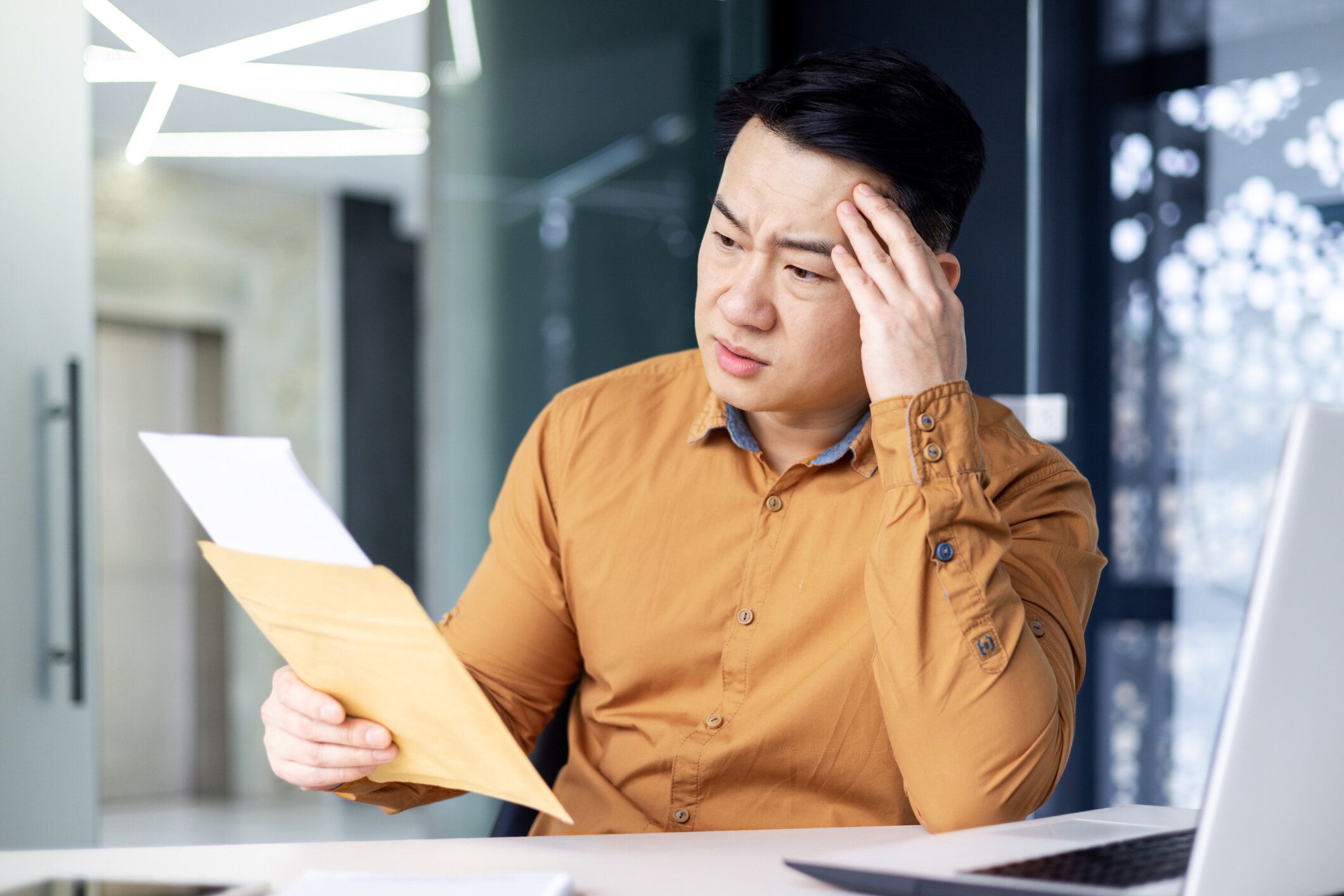 悪いニュースで手紙を読む真面目で動揺したアジアのビジネスマン、ノートパソコンを使って現代のオフィスビルで働く投資家。