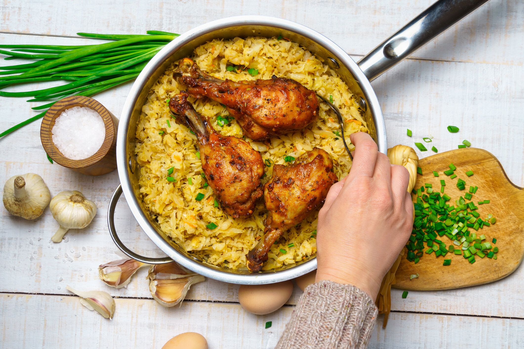 女性の手は、フライド刻みキャベツ、ニンニクとネギ、チーズ、農場の卵と塩、白い木製のボード上の素朴なスタイルで調理された食べ物の完全なフライパンにフライドチキン肉を置きます