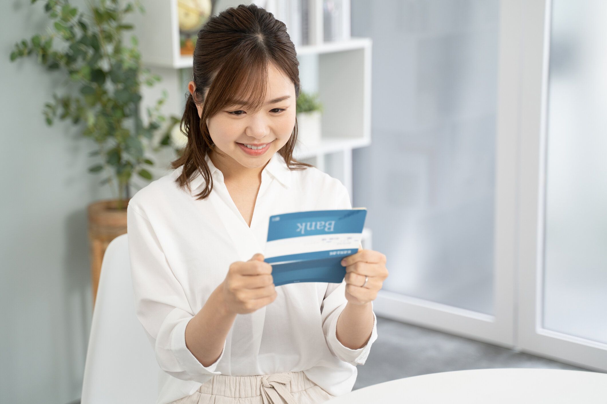 貯蓄通帳を手にした微笑む女性。女性が手にしている帳簿は、架空の日本の銀行の名前が書かれた通帳。